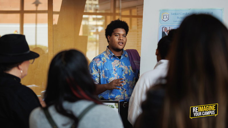 A person in a blue floral shirt presenting to a group of people in a room with posters on the wall
