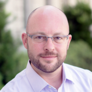 Headshot of white man with glasses and a beard. Wearing a light purple shirt