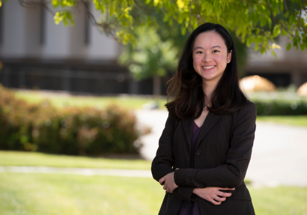 Female asian student with black suit. She is crossing her arms and has greenery in the background