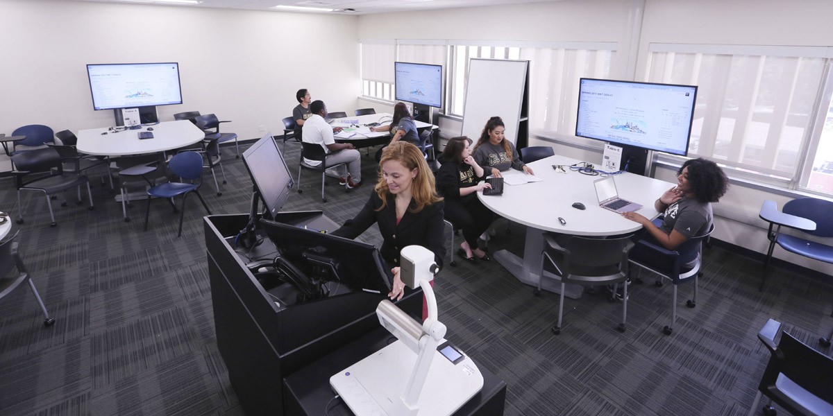 A modern classroom with computer stations and a professor giving a lecture through the available technology.