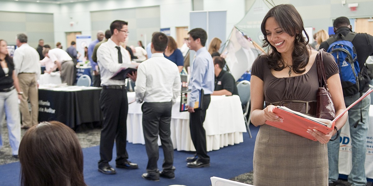 A student standing and smiling while holding an open binder and looking down at a person seated at a table.