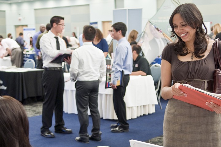A student standing and smiling while holding an open binder and looking down at a person seated at a table. 